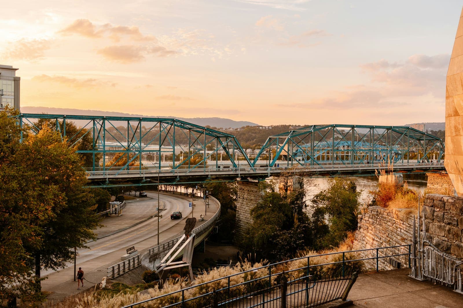 The Walnut Street Bridge over the Tennessee River in downtown Chattanooga at sunset, with the river winding through wooded hills toward Lookout Mountain in the distance.